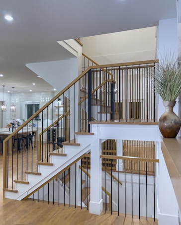 A modern interior with a staircase featuring wooden steps and black metal railings. The space is well-lit with recessed lighting and pendant lights. The walls are painted white, and the flooring is a light wood finish. A large ceramic vase with decorative grass is placed on a shelf. In the background, there is a kitchen area with bar stools and various kitchen appliances.