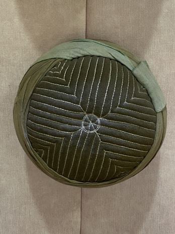 A round, textured cushion with a quilted green fabric exterior. It features concentric stitching patterns and is placed vertically against a plain fabric wall.