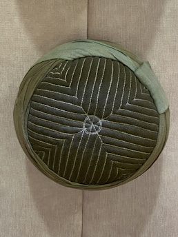 A round, textured cushion with a quilted green fabric exterior. It features concentric stitching patterns and is placed vertically against a plain fabric wall.