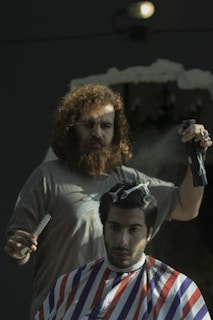 A man with curly hair and a beard is standing behind another man seated in a barber chair, trimming his hair. The seated man has his hair partially clipped and is draped with a barber cape featuring red, white, and blue stripes. The barber is holding a pair of scissors and a spray bottle.