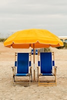 Two blue beach chairs with wooden frames are placed side by side on sandy ground. Above them, a large yellow umbrella provides shade. The background features a beach setting with overcast skies and some vegetation.