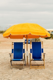 Two blue beach chairs with wooden frames are placed side by side on sandy ground. Above them, a large yellow umbrella provides shade. The background features a beach setting with overcast skies and some vegetation.