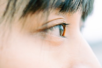 A close-up side view of a person's eye with focus on the eyelashes and eyebrow. The background is blurred, emphasizing the details of the eye and surrounding skin.