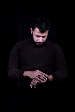 A man is seated in a dark setting, looking down with his hands crossed. He is dressed in a dark sweater, and the scene is largely monochromatic with a focus on his contemplative pose.