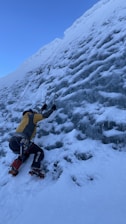 A person is ice climbing on a steep, frozen wall using ice axes and crampons. The climber is wearing climbing gear including a helmet, harness, and outdoor clothing. Snow and ice cover the surface, and a rope is attached for safety.