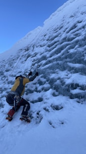 A person is ice climbing on a steep, frozen wall using ice axes and crampons. The climber is wearing climbing gear including a helmet, harness, and outdoor clothing. Snow and ice cover the surface, and a rope is attached for safety.