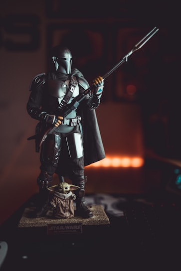 A detailed figurine of a sci-fi character in armor holding a weapon is displayed on a base. Another character, resembling a small alien with large ears, is at the foot of the larger figure. The lighting is dramatic, with warm tones highlighting the figures against a dark background.