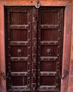 A robust wooden double door with metal studs and intricate carvings, framed by a reddish-brown stone. Ornate metal handles are positioned in the center of each door, and a chain with a padlock hangs at the top.