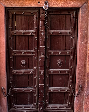 A robust wooden double door with metal studs and intricate carvings, framed by a reddish-brown stone. Ornate metal handles are positioned in the center of each door, and a chain with a padlock hangs at the top.