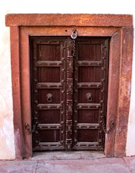 A rustic, wooden double door with intricate carvings set within a stone frame. The door is dark brown with decorative iron handles and studs. A chain and padlock suggest it is meant to be locked, possibly leading to a private or secure space. The surrounding wall is made of stone with a slightly textured appearance.