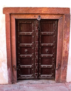 A rustic, wooden double door with intricate carvings set within a stone frame. The door is dark brown with decorative iron handles and studs. A chain and padlock suggest it is meant to be locked, possibly leading to a private or secure space. The surrounding wall is made of stone with a slightly textured appearance.