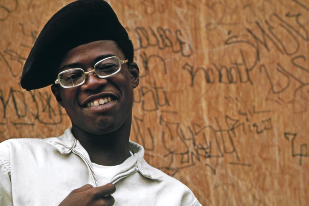 a man wearing glasses and a hat standing in front of a wooden wall