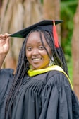 A person smiles while wearing a black graduation gown with a yellow sash, and holds the edge of a mortarboard cap adorned with a red tassel. The background features blurred wooden elements and greenery.