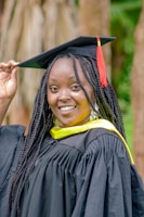 A person smiles while wearing a black graduation gown with a yellow sash, and holds the edge of a mortarboard cap adorned with a red tassel. The background features blurred wooden elements and greenery.