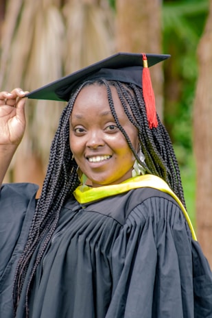 A person smiles while wearing a black graduation gown with a yellow sash, and holds the edge of a mortarboard cap adorned with a red tassel. The background features blurred wooden elements and greenery.