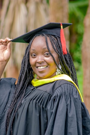 A person smiles while wearing a black graduation gown with a yellow sash, and holds the edge of a mortarboard cap adorned with a red tassel. The background features blurred wooden elements and greenery.