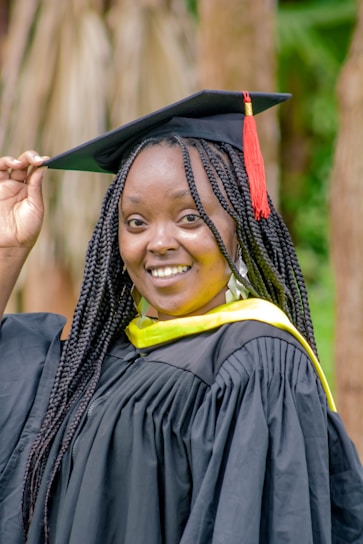 A person smiles while wearing a black graduation gown with a yellow sash, and holds the edge of a mortarboard cap adorned with a red tassel. The background features blurred wooden elements and greenery.
