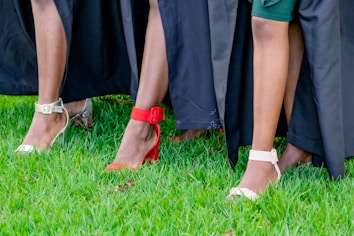 A close-up view of people wearing stylish shoes, featuring various high heels in red, white, and silver against a vibrant green grass background with black fabric draped around.