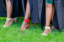 A close-up view of people wearing stylish shoes, featuring various high heels in red, white, and silver against a vibrant green grass background with black fabric draped around.