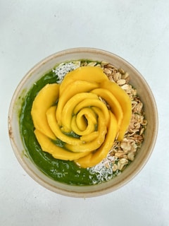 A smoothie bowl topped with a rose-shaped arrangement of mango slices, accompanied by granola and shredded coconut. The smoothie appears to be green in color and the bowl is placed on a white surface.