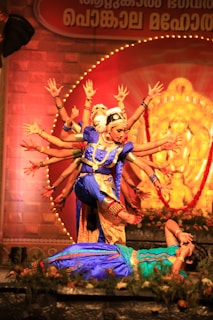 A traditional dance performance with dancers in vibrant blue and gold costumes. The central figure, wearing ornate jewelry, is in an intricate pose, surrounded by others extending their arms in a pattern. The background features a large illuminated figure and decorative elements.