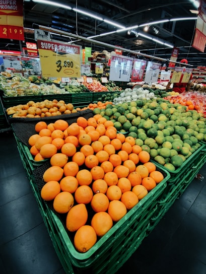 A supermarket display with large stacks of fresh oranges, limes, and other citrus fruits arranged neatly in green crates. Prominent signage displaying prices and promotional offers hangs above the produce section. The environment is well-lit with overhead fluorescent lights reflecting off the polished floor.