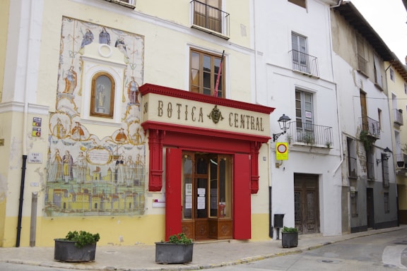 An old building with a red-framed entrance labeled 'Botica Central' on a narrow street features a large decorative mural with historic themes and religious figures. The facade is adorned with traditional architecture, including balconies and street lamps. Small potted plants are placed by the entrance.