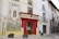 An old building with a red-framed entrance labeled 'Botica Central' on a narrow street features a large decorative mural with historic themes and religious figures. The facade is adorned with traditional architecture, including balconies and street lamps. Small potted plants are placed by the entrance.