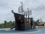 A playground structure designed to resemble a pirate ship with sails and an anchor emblem prominently displayed. It is set on a blue and tan rubberized surface with trees and a building visible in the background under a cloudy sky.