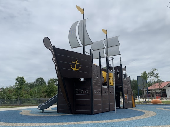 A playground structure designed to resemble a pirate ship with sails and an anchor emblem prominently displayed. It is set on a blue and tan rubberized surface with trees and a building visible in the background under a cloudy sky.