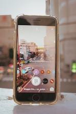 A smartphone on a white surface displays a cityscape with traffic on the screen. The device appears to be using a photo editing or streaming app, with an interface overlaid on the image showing several rounded buttons with numbers and icons. In the background, an out-of-focus view of city buildings and roads adds depth to the composition.