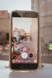 A smartphone on a white surface displays a cityscape with traffic on the screen. The device appears to be using a photo editing or streaming app, with an interface overlaid on the image showing several rounded buttons with numbers and icons. In the background, an out-of-focus view of city buildings and roads adds depth to the composition.