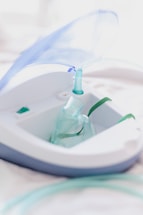 A close-up view of a nebulizer device with a transparent mask and tubing. The machine has a sleek design with white and gray colors, and a blue lid is slightly opened. The focus is on the medical equipment, highlighting its purpose for respiratory treatment.