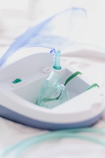 A close-up view of a nebulizer device with a transparent mask and tubing. The machine has a sleek design with white and gray colors, and a blue lid is slightly opened. The focus is on the medical equipment, highlighting its purpose for respiratory treatment.