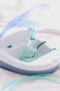A close-up view of a nebulizer device with a transparent mask and tubing. The machine has a sleek design with white and gray colors, and a blue lid is slightly opened. The focus is on the medical equipment, highlighting its purpose for respiratory treatment.