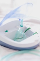 A close-up view of a nebulizer device with a transparent mask and tubing. The machine has a sleek design with white and gray colors, and a blue lid is slightly opened. The focus is on the medical equipment, highlighting its purpose for respiratory treatment.