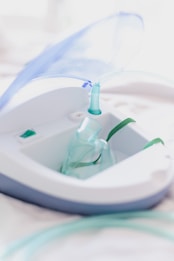 A close-up view of a nebulizer device with a transparent mask and tubing. The machine has a sleek design with white and gray colors, and a blue lid is slightly opened. The focus is on the medical equipment, highlighting its purpose for respiratory treatment.