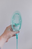A hand is holding a transparent oxygen mask against a plain wall. The mask is attached to a green tube, and the hand appears slightly tattooed or marked. The overall lighting is soft, creating a clinical yet calm atmosphere.