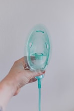 A hand is holding a transparent oxygen mask against a plain wall. The mask is attached to a green tube, and the hand appears slightly tattooed or marked. The overall lighting is soft, creating a clinical yet calm atmosphere.