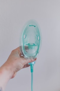 A hand is holding a transparent oxygen mask against a plain wall. The mask is attached to a green tube, and the hand appears slightly tattooed or marked. The overall lighting is soft, creating a clinical yet calm atmosphere.