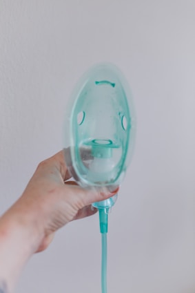 A hand is holding a transparent oxygen mask against a plain wall. The mask is attached to a green tube, and the hand appears slightly tattooed or marked. The overall lighting is soft, creating a clinical yet calm atmosphere.
