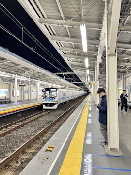 A modern train is arriving at a platform under bright fluorescent lighting. The station is clean and well-maintained, with a few passengers waiting on the platform. Overhead power lines run above the tracks, and there are clear markings and signs along the platform edge.