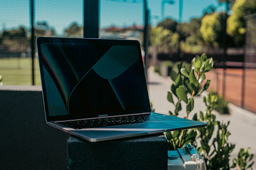 A Trustpoint laptop open on a park bench, surrounded by green leaves and sunlight filtering through trees.