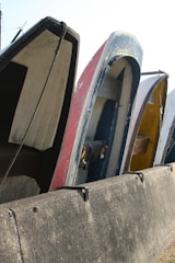 Several boats are placed vertically against a concrete barrier, with visible wear and paint peeling from their surfaces. The boats are lined up closely, showcasing various colors and the ropes hanging loosely.