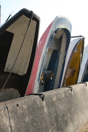 Several boats are placed vertically against a concrete barrier, with visible wear and paint peeling from their surfaces. The boats are lined up closely, showcasing various colors and the ropes hanging loosely.