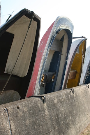 Several boats are placed vertically against a concrete barrier, with visible wear and paint peeling from their surfaces. The boats are lined up closely, showcasing various colors and the ropes hanging loosely.
