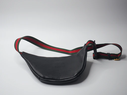 A black leather waist bag with a contrasting strap featuring red and green stripes is shown. The strap includes an adjustable buckle and a gold metal clip. The bag is laid out on a plain light gray surface, creating a minimalistic backdrop.