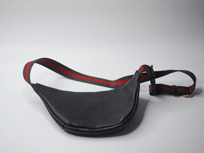 A black leather waist bag with a contrasting strap featuring red and green stripes is shown. The strap includes an adjustable buckle and a gold metal clip. The bag is laid out on a plain light gray surface, creating a minimalistic backdrop.