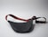 A black leather waist bag with a contrasting strap featuring red and green stripes is shown. The strap includes an adjustable buckle and a gold metal clip. The bag is laid out on a plain light gray surface, creating a minimalistic backdrop.