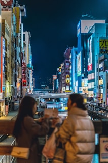 A vibrant urban night scene featuring a bustling canal lined with illuminated buildings displaying colorful signs. Two people in the foreground appear to be in conversation, adding a sense of lively interaction to the setting.
